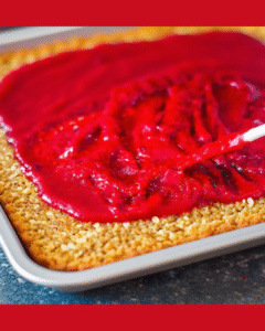 Cranberry jam being spread evenly across the oat and coconut crust layer to form the base of the vegan dessert bars.