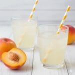 A refreshing glass of peachy lemonade with ice cubes and fresh peach slices, set on a sun-drenched wooden surface with soft garden greenery in the background.
