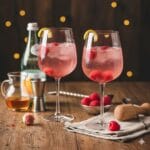 Two elegant wine glasses filled with a sparkling pink raspberry rose spritz, garnished with fresh raspberries and lemon twists, set on a rustic wooden table alongside a cocktail jigger, muddler, and fresh berries.