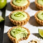 A top-down overhead shot of elegant mini vegan key lime cheesecake bites arranged on a white marble serving platter, each bite featuring a pale green creamy cashew-lime filling with a vibrant lime-yellow hue, sitting atop golden-brown graham-style coconut crusts in individual fluted mini tart pans, garnished with delicate curls of fresh lime zest on top of each bite, one bite cut in half to reveal the distinct layers of buttery coconut crust and silky smooth lime filling, natural daylight streaming from the side creating soft shadows, scattered fresh key limes and lime wedges around the platter, a few loose cashews and shredded coconut artfully placed nearby, shot with shallow depth of field focusing on the center bites while the background gently blurs, professional food photography styling with a clean minimalist aesthetic, crisp details showing the creamy texture of the filling and the crumbly crust
