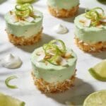 A overhead flat-lay photograph of mini tropical coconut-lime vegan cheesecake bites arranged on a white marble surface, each bite showcasing a golden toasted coconut crust base topped with a pale green creamy cashew-lime filling, garnished with fresh lime zest curls and toasted coconut flakes, some lime wedges and halved limes scattered artfully around the bites, drops of coconut cream pooled decoratively on the marble, natural window lighting creating soft shadows, professional food styling with a light and airy tropical aesthetic, shallow depth of field with the front cheesecake bites in sharp focus showing the distinct layers of toasted coconut crust and vibrant lime filling