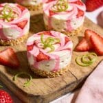 A professional overhead food photography shot of mini vegan key lime cheesecake bites with vibrant pink strawberry swirls marbled through creamy white cashew filling, displayed on a rustic wooden serving board, each bite-sized dessert nestled in a golden coconut graham crust, garnished with fresh lime zest curls and thin strawberry slices, surrounded by whole fresh strawberries and lime wedges, natural daylight streaming from the side creating soft shadows, shallow depth of field focusing on the front cheesecake bites revealing the beautiful pink and white swirl pattern, pastel linen napkin artfully placed beside the board, professional food styling with a bright, airy, summery aesthetic
