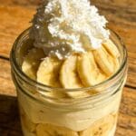 Overhead shot of elegant glass dessert jar layered with ivory banana cream custard, fresh ripe banana slices, and fluffy whipped coconut topping, golden natural light, rustic wood table, cozy bakery presentation